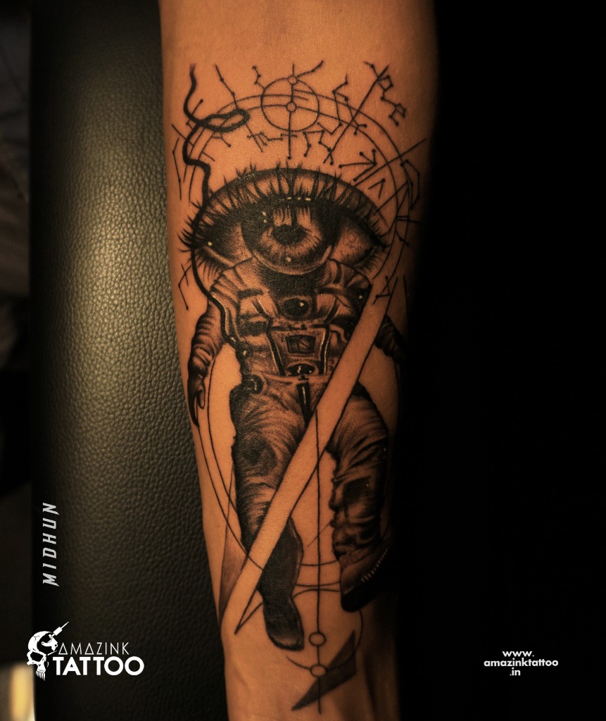 Surrealist astronaut blackwork forearm tattoo with a large all-seeing eye replacing the helmet visor, surrounded by celestial and geometric alignments, custom designed at Amazing Tattoo in Kozhikode.