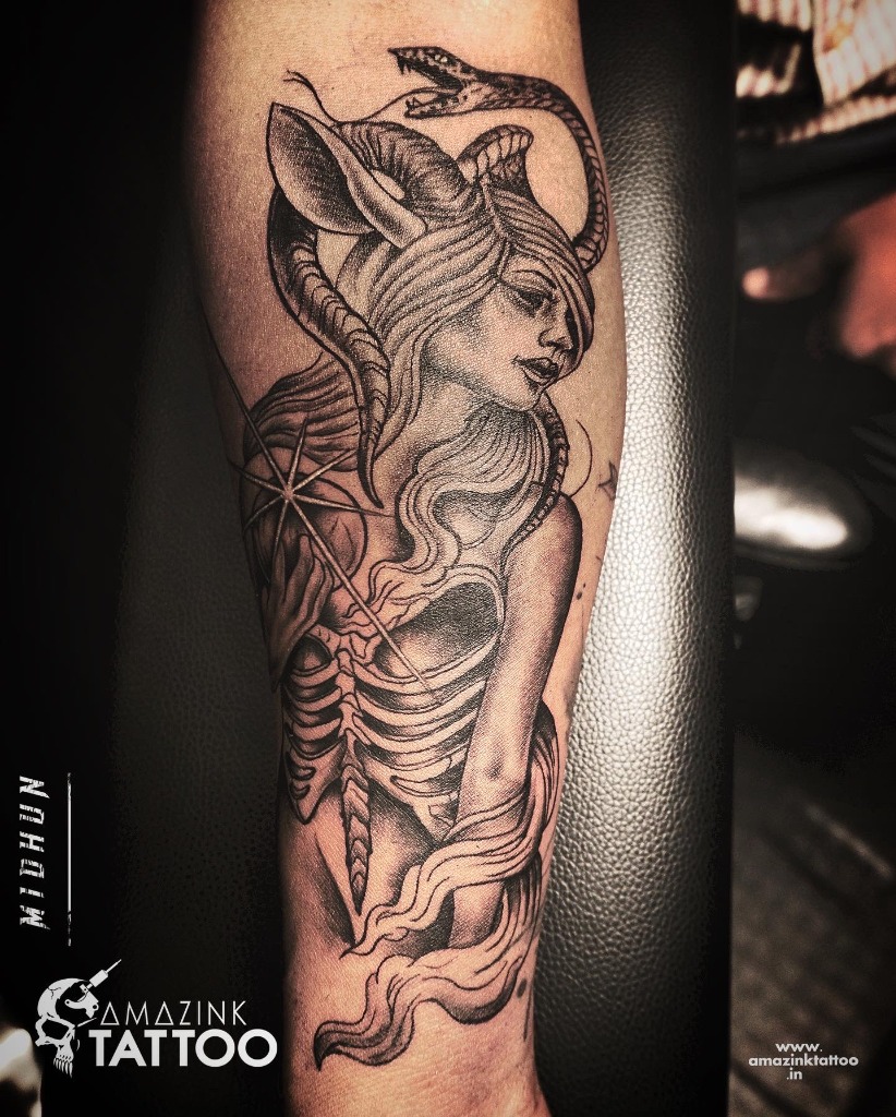 A surreal blackwork forearm tattoo of a mystical horned goddess with a coiled snake and an exposed skeletal ribcage, custom designed by Midhun at Amazing Tattoo in Kerala.