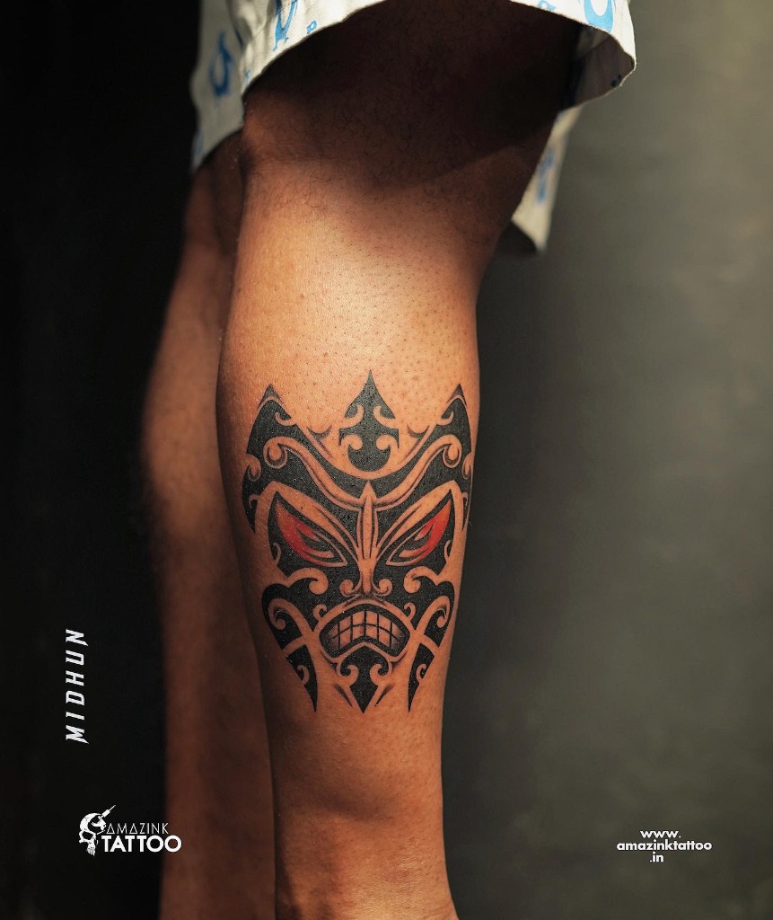 Bold Polynesian tribal mask leg tattoo on the calf with sharp blackwork curls and striking red accent eyes, inked by Midhun at Amazing Tattoo in Kozhikode.